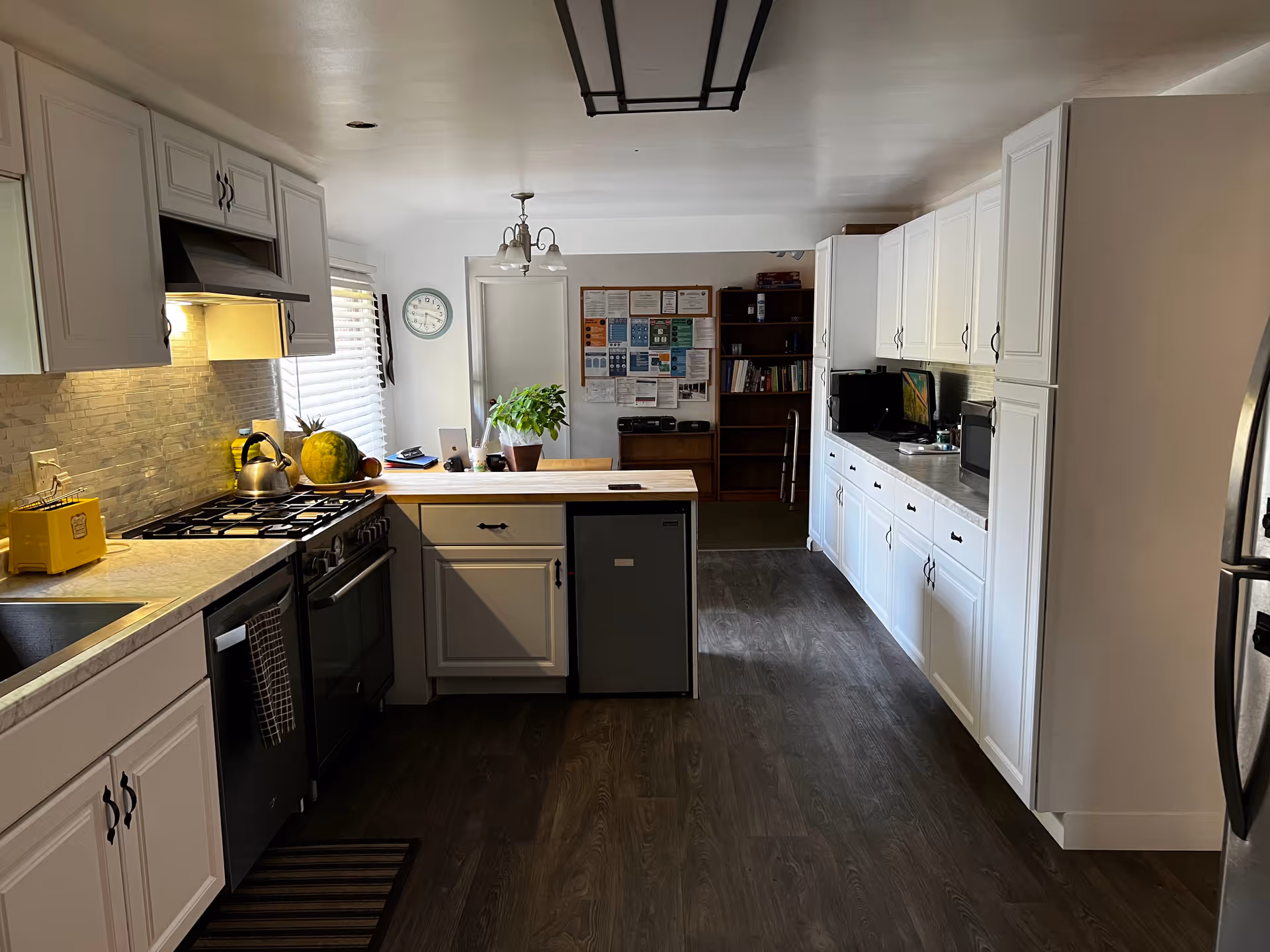 A bright kitchen with white cabinets, a stove with a kettle, a yellow toaster, a small refrigerator under a wooden countertop, and a microwave on the far counter. There is a plant on the countertop and a clock on the wall near a door. The floor is dark wood, and there is a bulletin board and bookshelf visible in the background.