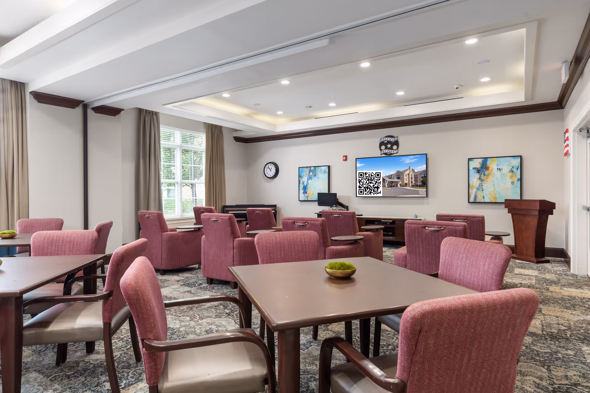 A well-lit common room with multiple tables and pink upholstered chairs arranged for group seating. The room features large windows with beige curtains, a patterned carpet, and a ceiling with recessed lighting. On the far wall, there is a large flat-screen TV displaying an image of a building, flanked by two abstract paintings. A clock and a wooden podium are also visible in the room.