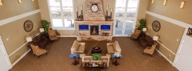 Spacious living room area with beige walls and carpeted floor, featuring two beige sofas and two brown armchairs arranged around a central coffee table. A stone fireplace with a clock and candle holders is centered on the far wall, flanked by two flat-screen TVs. Large windows and glass doors allow natural light to fill the room, and decorative plants and wall art add to the cozy atmosphere.