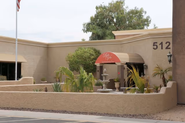Exterior view of Bella Vita Health & Rehabilitation Center showing a beige stucco building with the number 512 on the wall, an American flag on a pole, a small garden with plants and a fountain, and a red awning above the entrance displaying the facility's name.