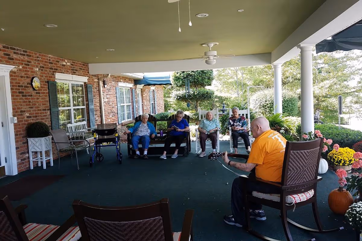 A covered outdoor patio area at a senior living facility where four elderly women are seated in chairs along a brick wall, and a man in an orange shirt is playing guitar facing them. The patio is decorated with potted flowers and pumpkins, with white columns and greenery visible outside.