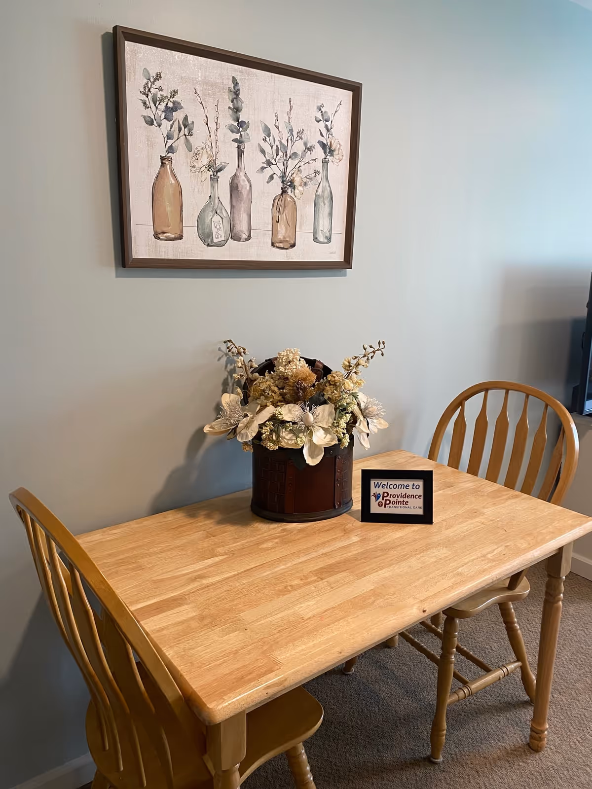 A small wooden dining table with two matching wooden chairs. On the table is a decorative flower arrangement in a brown container and a small framed sign that reads 'Welcome to Providence Pointe Transitional Care.' Above the table hangs a framed artwork depicting five glass bottles with flowers inside against a light-colored wall.