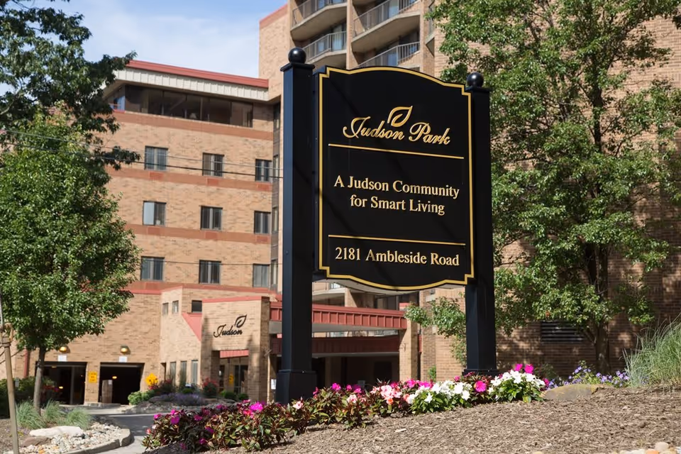 Outdoor view of Judson Park senior living community sign with the building in the background, surrounded by trees and flowers.