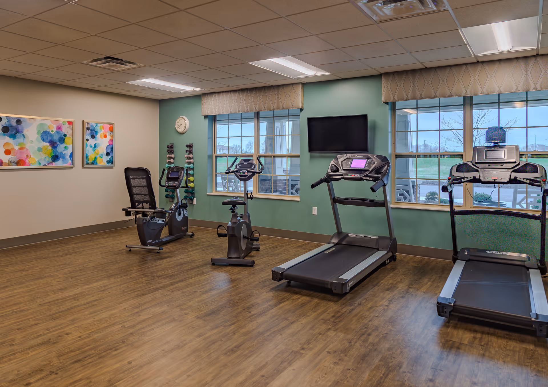 A senior living facility exercise room with two treadmills, two stationary bikes, a rack of colorful dumbbells, a wall clock, two large windows with beige valances, a wall-mounted flat screen TV, and two colorful abstract paintings on the wall. The room has wood flooring and a green accent wall.