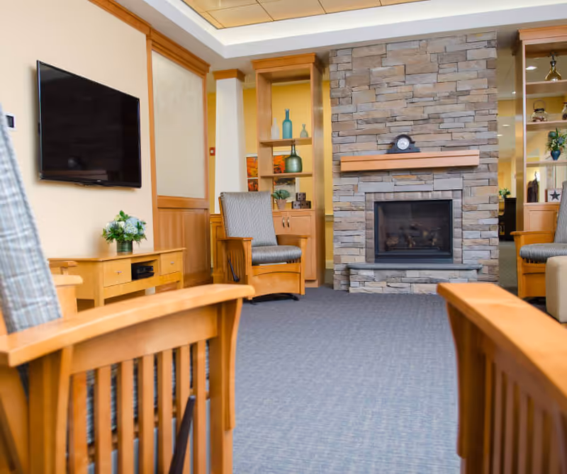A cozy living room area with wooden furniture including chairs and a TV stand, a flat-screen TV mounted on the wall, a stone fireplace with a clock on the mantel, and built-in shelves with decorative items.