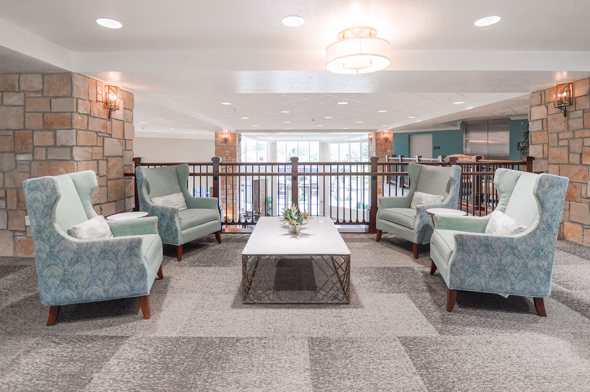 A seating area in a senior living facility with four light blue upholstered armchairs arranged around a rectangular coffee table with a small plant centerpiece. The area has stone pillars, carpeted flooring, and a railing overlooking a lower level with large windows in the background.