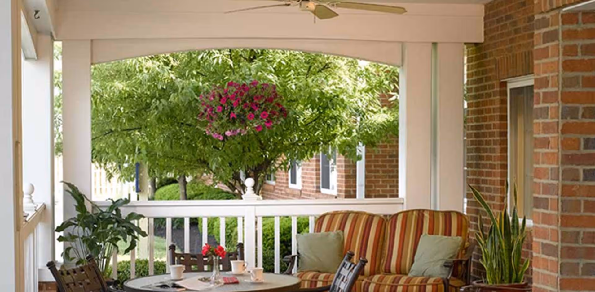 Covered porch with striped cushioned chairs and a round table set, hanging flowers and greenery beyond a white railing.