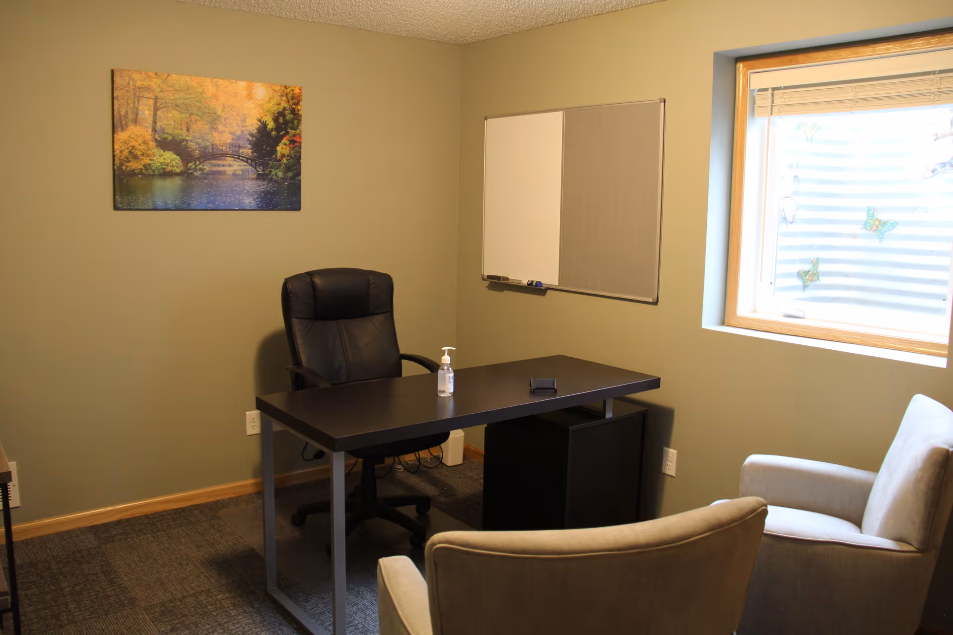 A small office room with a black desk and black office chair behind it. Two beige armchairs face the desk. On the desk is a bottle of hand sanitizer and a small black object. A window with blinds and butterfly decorations is on the right wall. A whiteboard and corkboard combination hangs on the wall behind the desk. A framed picture of a scenic autumn landscape with a bridge over water is on the left wall.