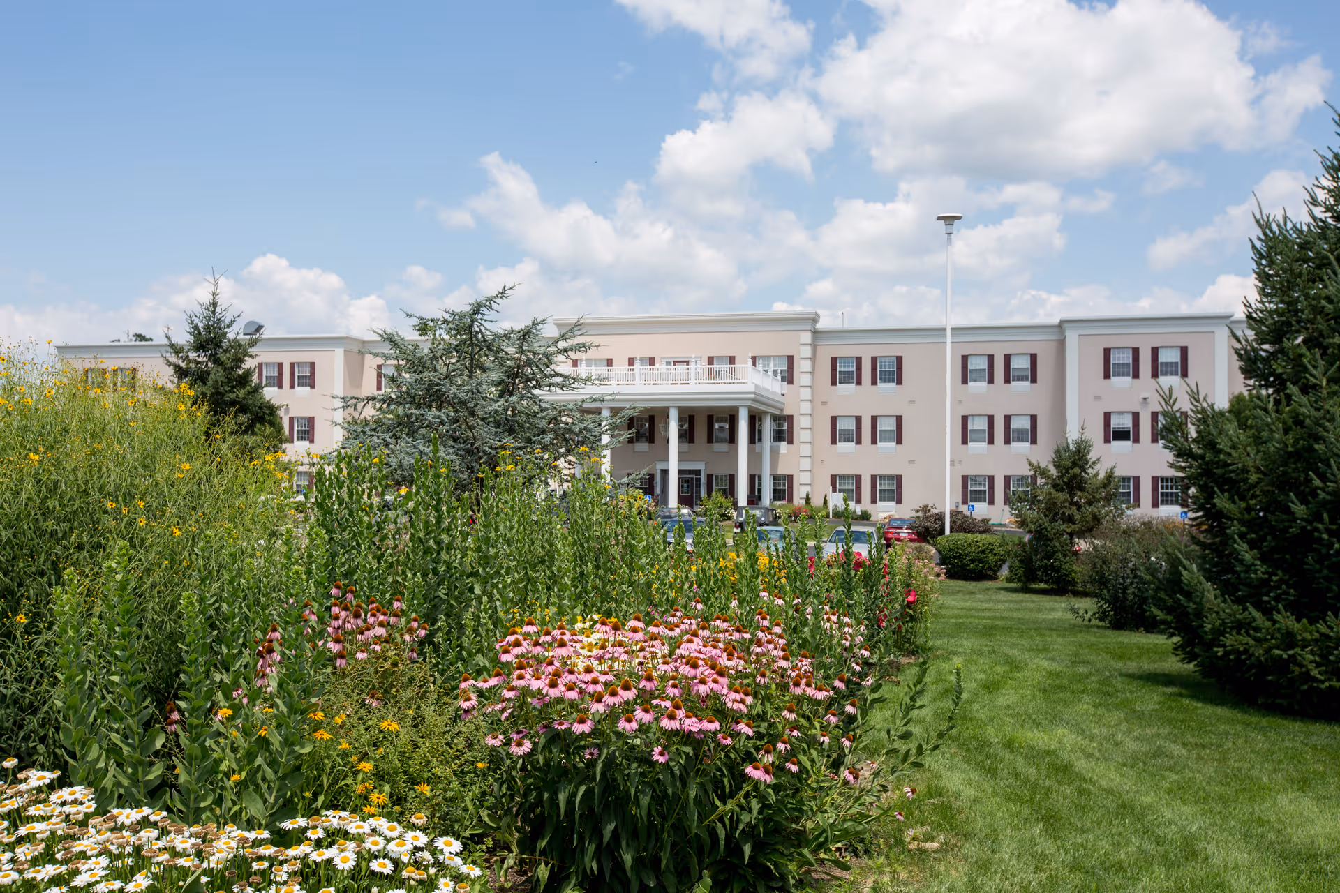 Exterior view of a three-story retirement community building with beige walls and maroon window shutters, surrounded by a lush garden with various flowers and green trees under a partly cloudy sky.