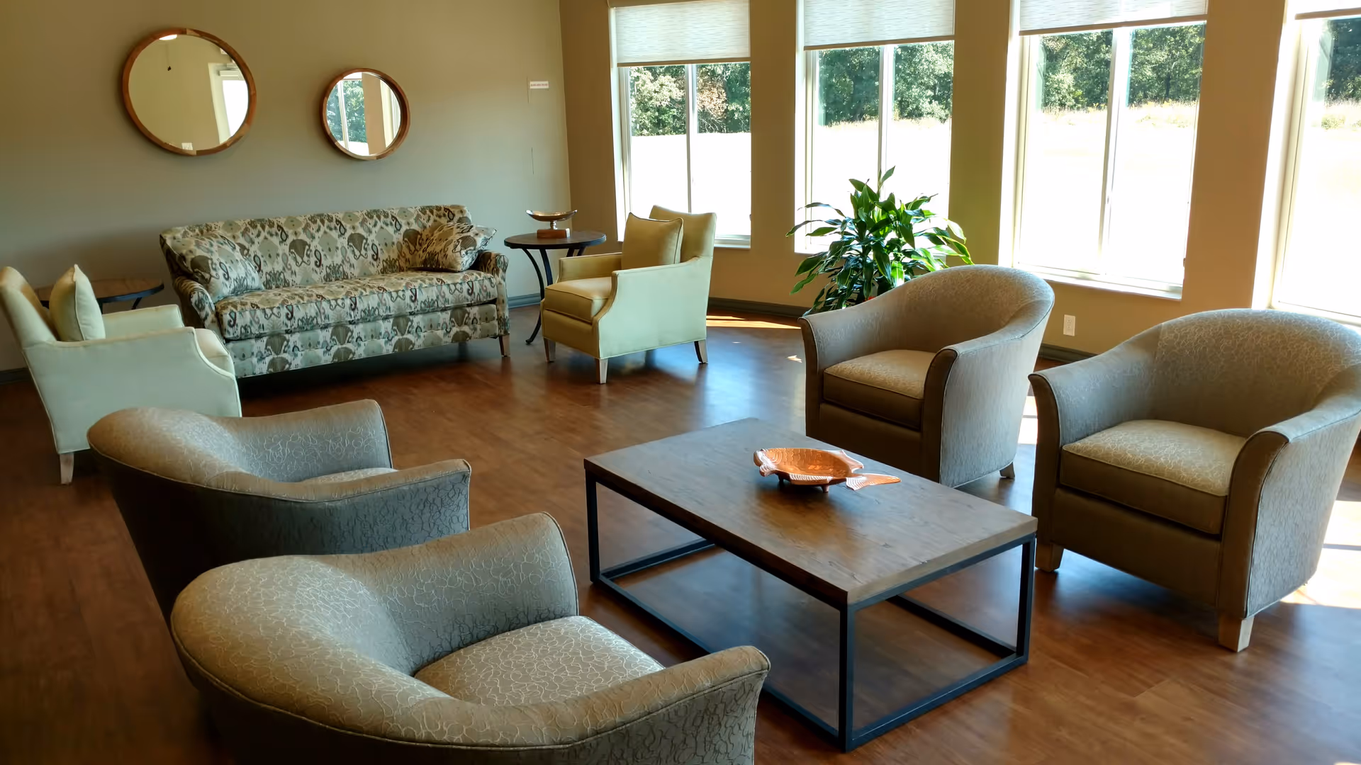 A bright and spacious living room with large windows letting in natural light. The room features a patterned sofa, several armchairs arranged around a rectangular wooden coffee table, two round mirrors on the wall, and a green potted plant near the windows. The floor is wooden and the walls are painted a neutral color.
