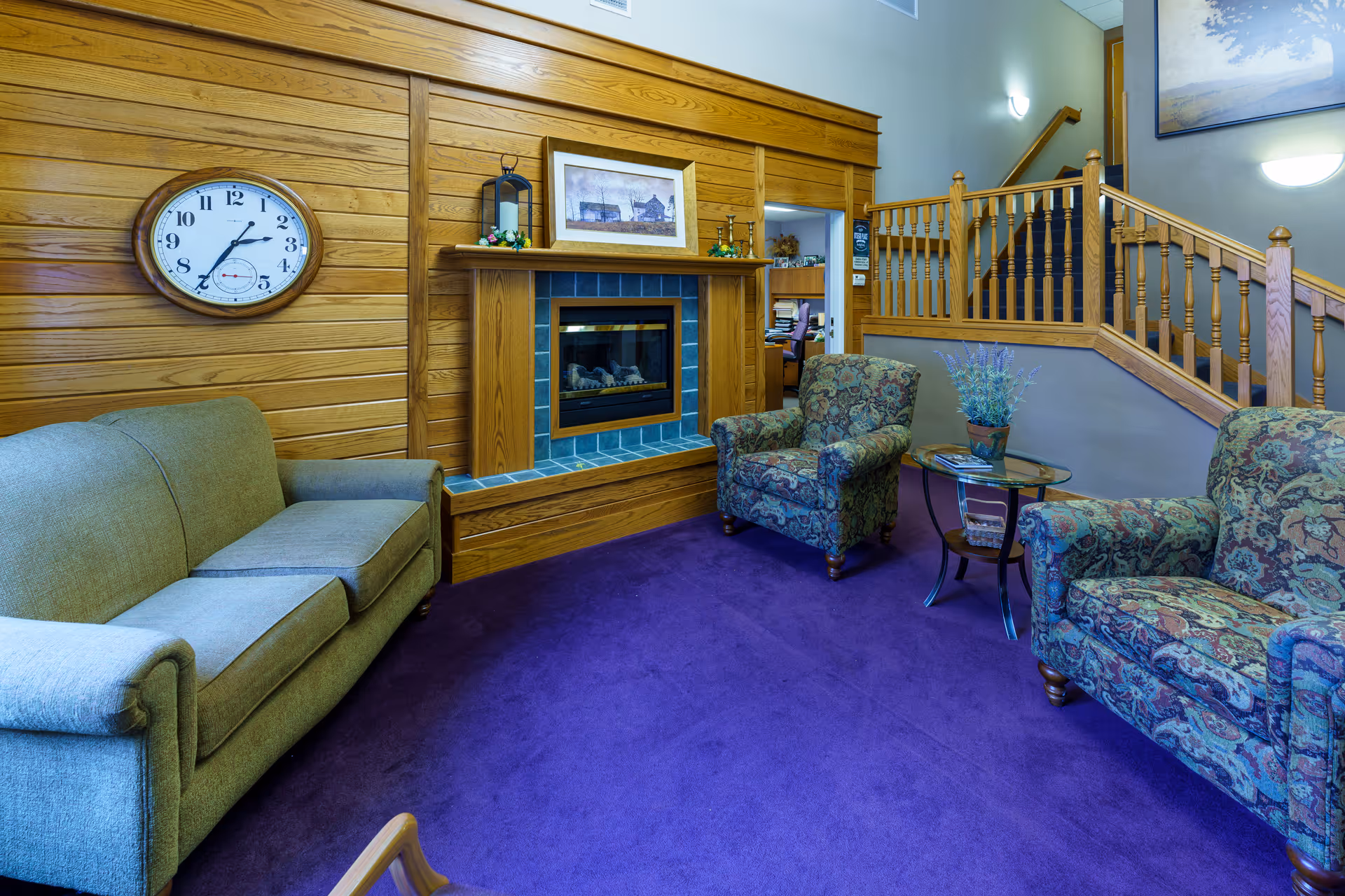 A cozy living room area with a green sofa and two patterned armchairs arranged around a glass-top side table with a potted plant. The room features a wooden paneled wall with a clock and a framed picture above a fireplace with blue tiles. A wooden staircase with a railing is visible in the background.