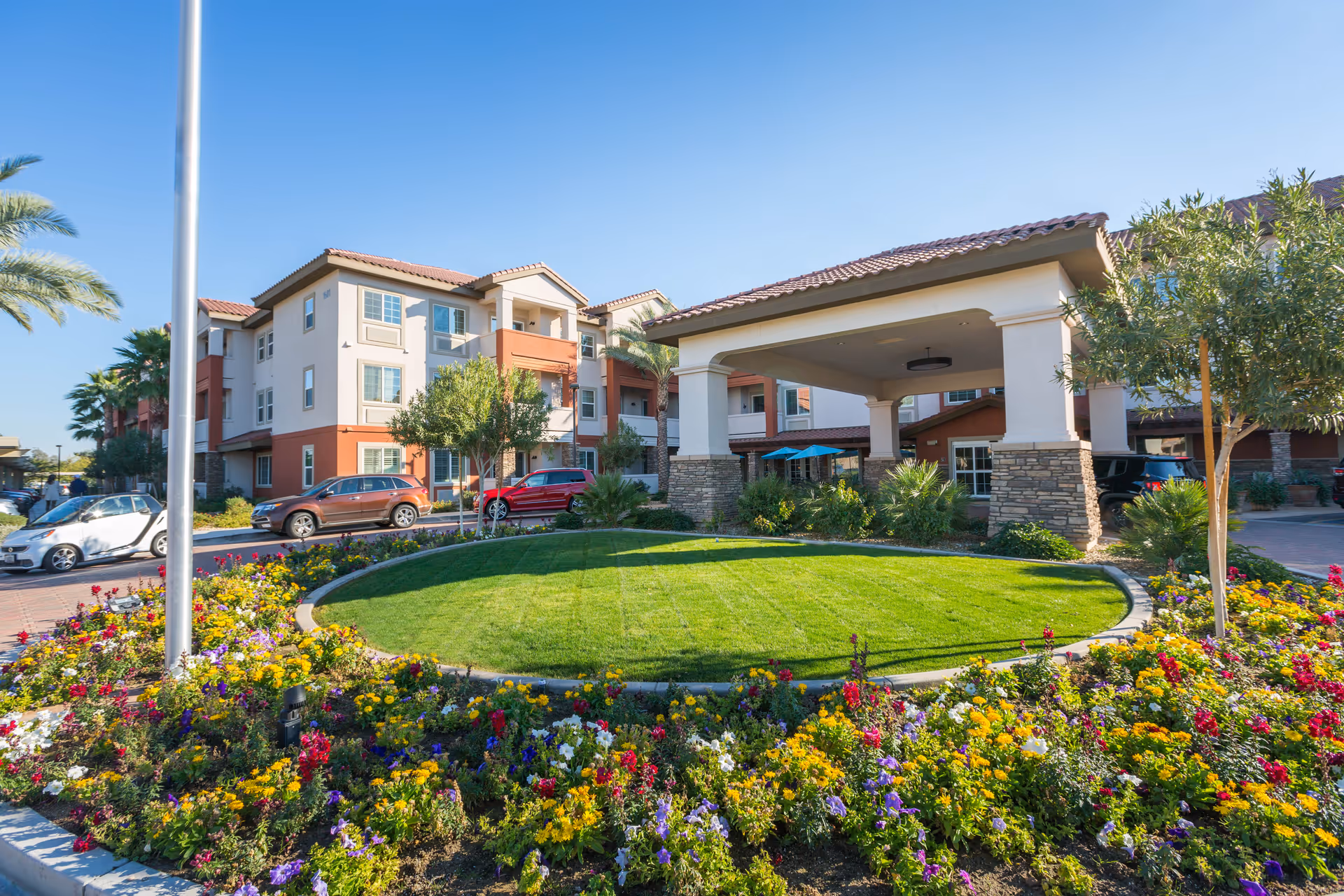 Exterior view of a senior living facility with a circular driveway surrounded by colorful flower beds and green grass. The building has a beige and terracotta color scheme with multiple windows and a covered entrance supported by stone pillars. Several cars are parked near the entrance under a clear blue sky.