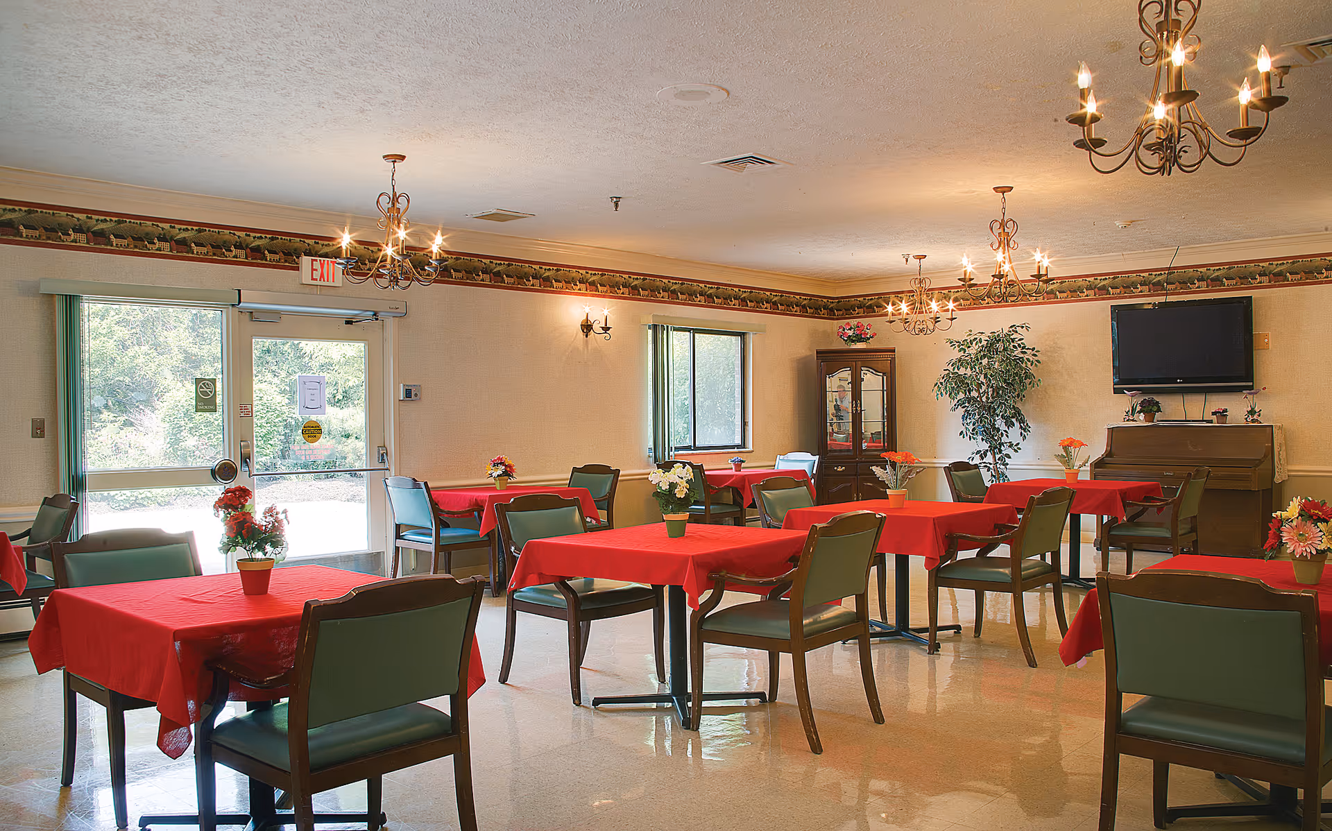 A dining room with multiple tables covered in red tablecloths, each table surrounded by green chairs. The room has chandeliers hanging from the ceiling, a large window, a glass door with an exit sign above it, a cabinet with glass doors, a piano with a TV mounted above it, and several potted plants and flower arrangements on the tables and around the room.