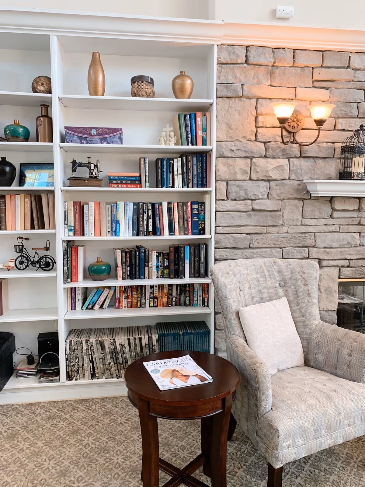 Cozy living room area with a beige patterned armchair with a white pillow, a small round wooden table with a magazine on it, a white built-in bookshelf filled with books and decorative items, and a stone wall with a double wall sconce light fixture and a white mantel holding a decorative birdcage.