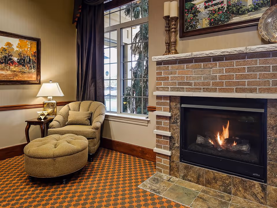 Cozy corner of a living room with a cushioned armchair and matching ottoman on a patterned carpet. A side table with a lamp and decorative items is next to the chair. A large window with dark curtains lets in natural light. A brick fireplace with a fire burning is on the right, topped with candles and a framed painting above it.