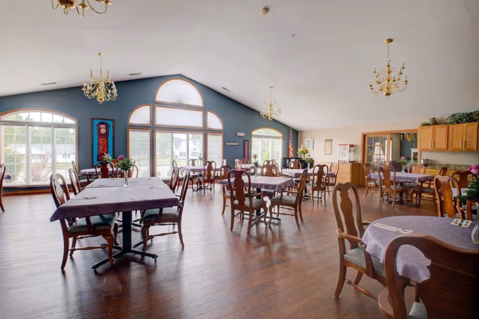 A spacious dining room with multiple wooden tables and chairs arranged neatly. Each table is covered with a purple tablecloth and has a small vase with flowers. Large windows with arched tops allow natural light to fill the room. The walls are painted blue and beige, and there are chandeliers hanging from the ceiling. Wooden cabinets and a door are visible in the background.