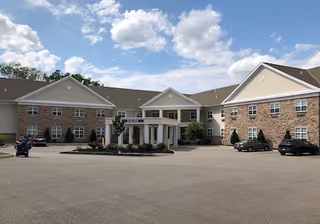 Front exterior view of a two-story health and rehab center building with a large driveway and several parked cars under a partly cloudy sky.