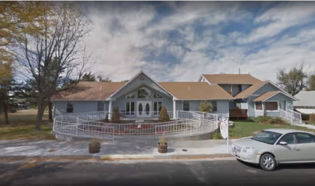 Front exterior view of a single-story assisted living facility with a ramp and stairs leading to the main entrance. The building has a light blue exterior with a brown roof, surrounded by trees and a parked silver car in front.