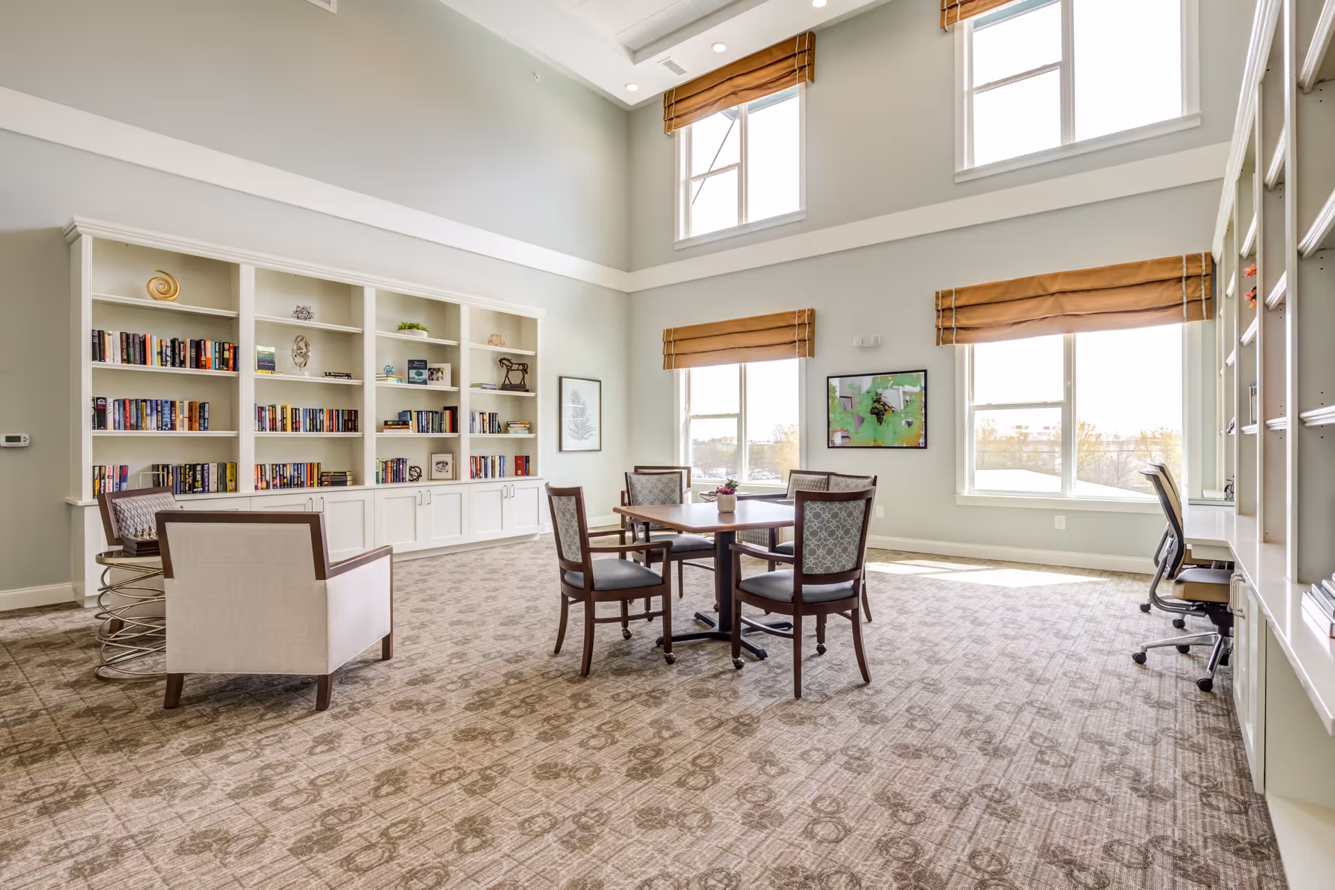 A bright and spacious common area with large windows letting in natural light. The room features a carpeted floor, a table with four chairs in the center, two armchairs, and built-in white bookshelves filled with books and decorative items. There are also two office chairs and a desk area along the right wall.
