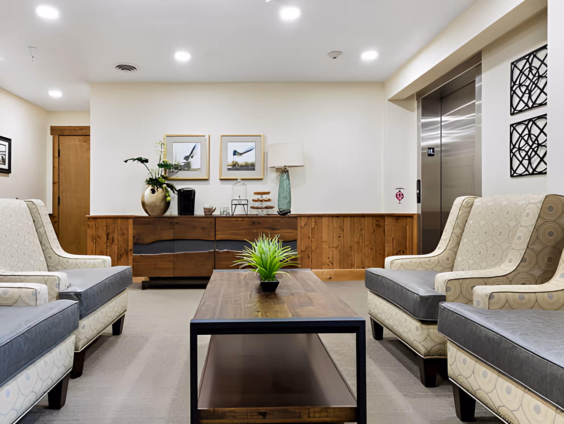 Lobby-style seating area with upholstered armchairs facing a wooden coffee table, a sideboard with decor, and an elevator in the background.