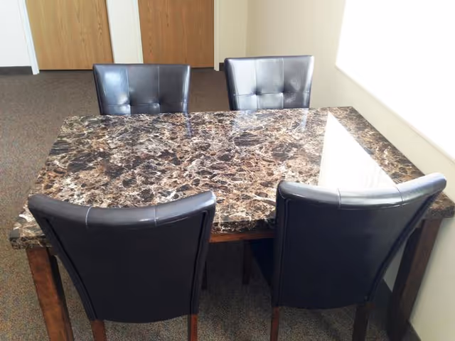 A rectangular dining table with a dark brown marble-like surface surrounded by four black cushioned chairs in a room with carpeted floor and wooden doors in the background.
