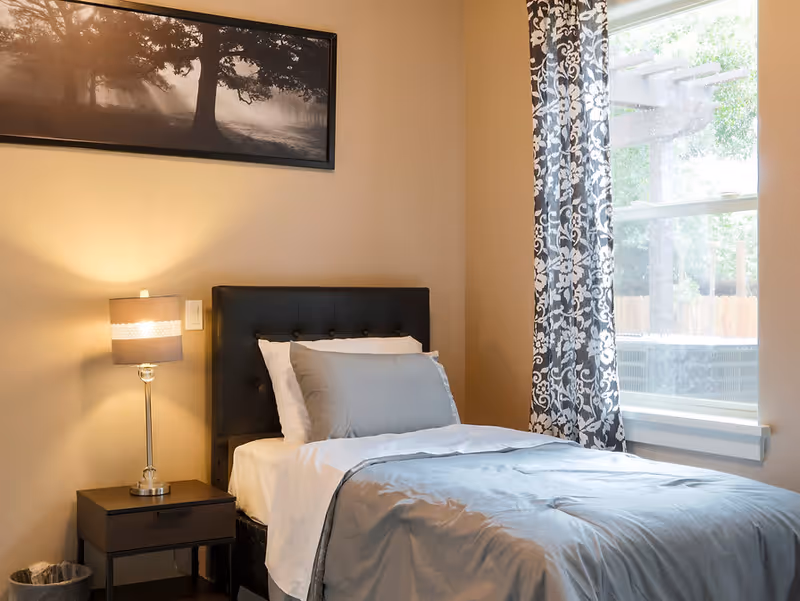 A neatly made single bed with gray and white bedding in a bedroom. Next to the bed is a dark wooden nightstand with a modern lamp emitting warm light. A framed black and white picture of a tree hangs on the beige wall above the bed. A window with patterned curtains allows natural light to enter the room.