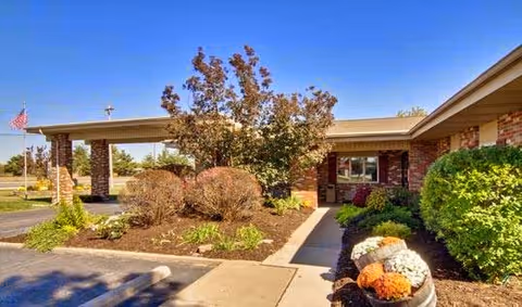 Entrance and landscaped front walkway of a single-story brick building with a covered drop-off and American flag.