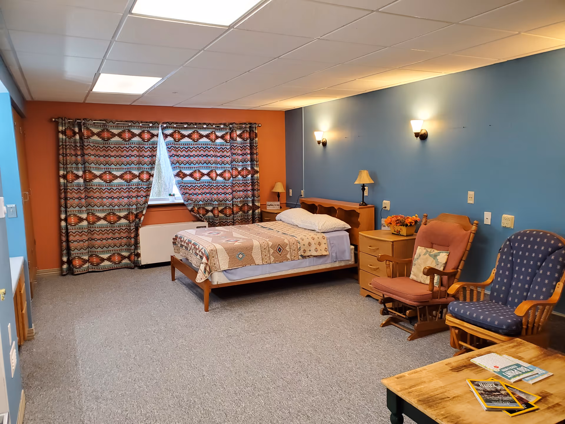 A furnished senior community bedroom with a single bed, nightstand, patterned curtains, and two upholstered rocking chairs.