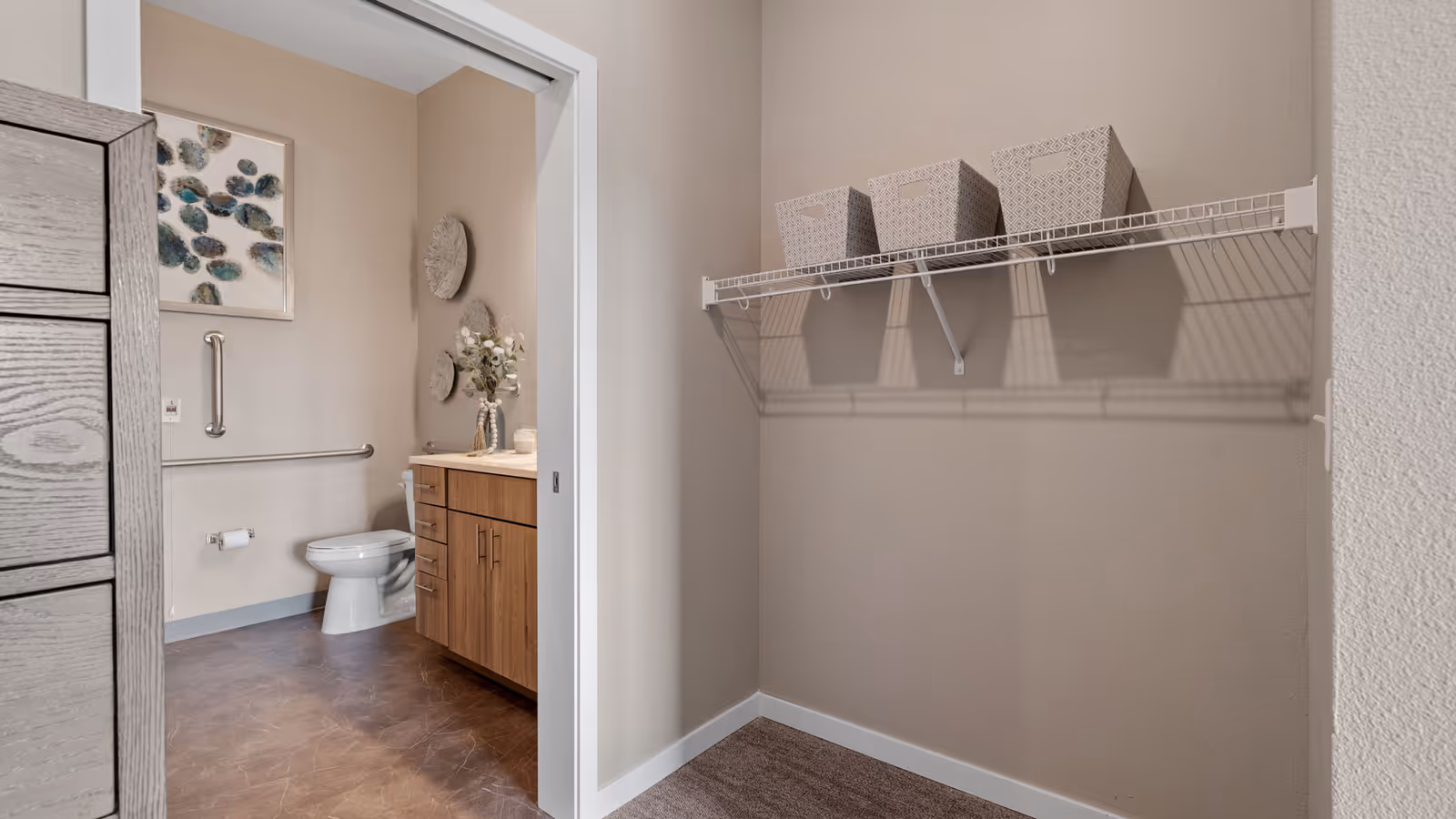 Interior view of an empty closet opening onto a bathroom with a toilet, wooden vanity, and wall decorations.
