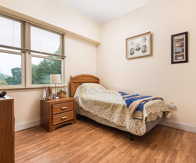 A small bedroom with a single bed covered in a floral quilt and a blue blanket. Next to the bed is a wooden nightstand with two drawers, a lamp, and decorative items. There is a large window with blinds partially open, showing greenery outside. The walls are light-colored with two framed pictures hanging above the bed. The floor is wooden.