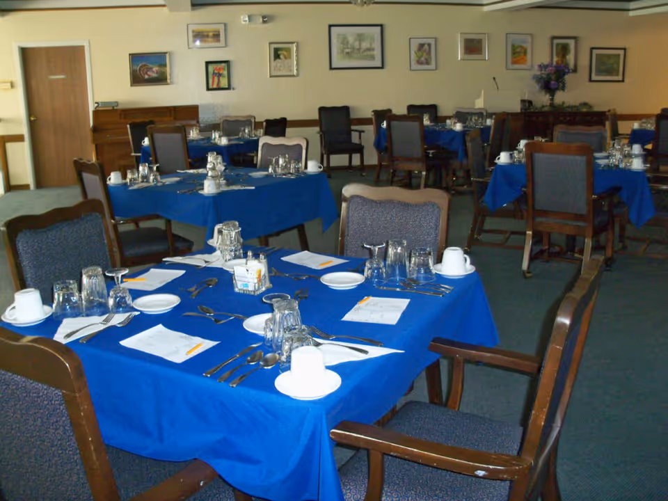 Dining room with multiple tables covered in blue tablecloths, set with plates, cups, glasses, silverware, and napkins. Chairs with wooden frames and upholstered seats surround the tables. Paintings and framed pictures hang on the beige walls, and a piano is visible in the background.
