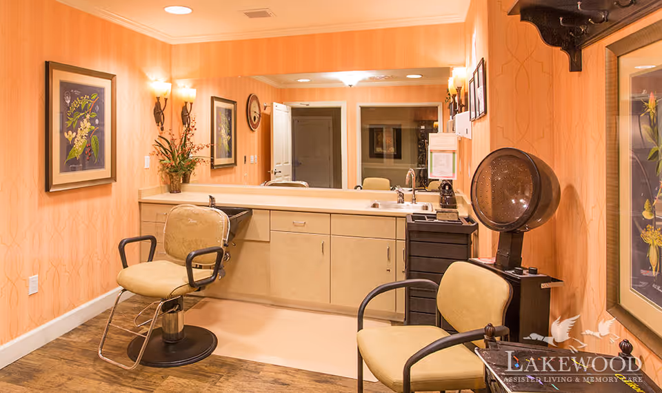 Salon-style grooming area with two styling chairs, a hair dryer station, sink and large mirror in a warm-toned interior.