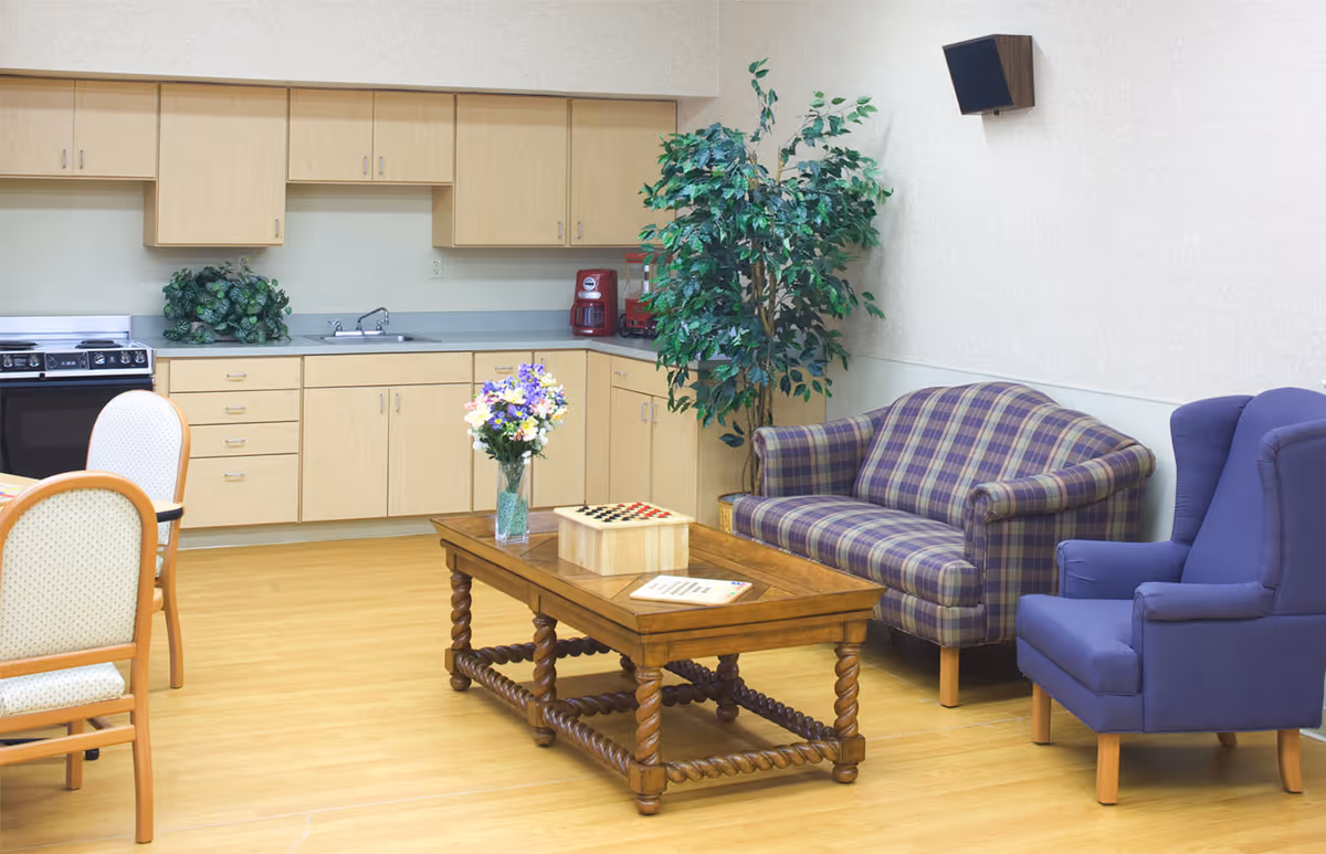 A cozy common area with a small kitchen in the background featuring light wood cabinets, a stove, sink, and coffee maker. In the foreground, there is a wooden coffee table with a vase of flowers and a checkers game on it. To the right, there is a plaid loveseat and a purple armchair, with a large potted plant behind the loveseat. The floor is light wood, and there is a wall-mounted speaker above the loveseat.