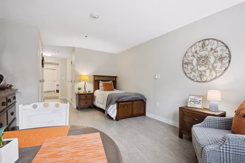 A neatly arranged bedroom in a senior living facility featuring a single bed with pillows and a blanket, a wooden nightstand with a lamp and a small plant, a decorative wall piece, a comfortable armchair with a cushion, and a small table with placemats. The room has light-colored walls and flooring, with a hallway visible in the background.