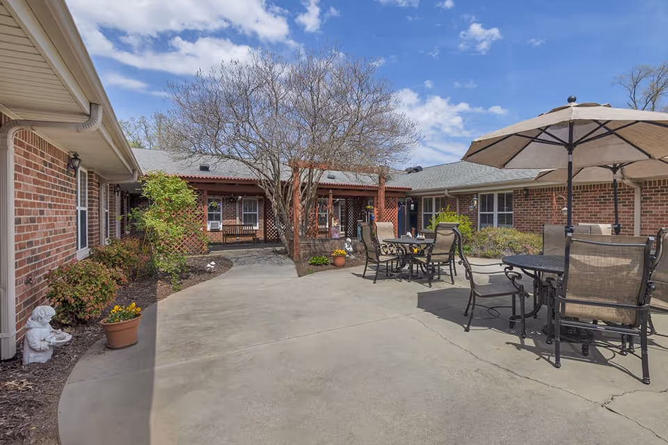 Outdoor courtyard with brick buildings, patio tables and umbrellas, potted plants, and a central tree.