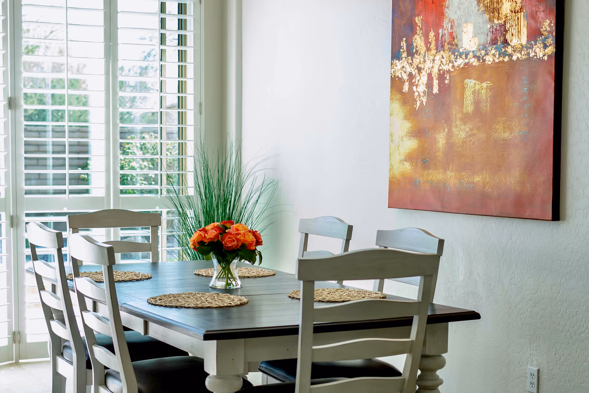 A dining area with a wooden table and six chairs. The table has woven placemats and a vase with orange roses in the center. There is a large window with white shutters letting in natural light, a tall green plant in the corner, and a colorful abstract painting on the wall.