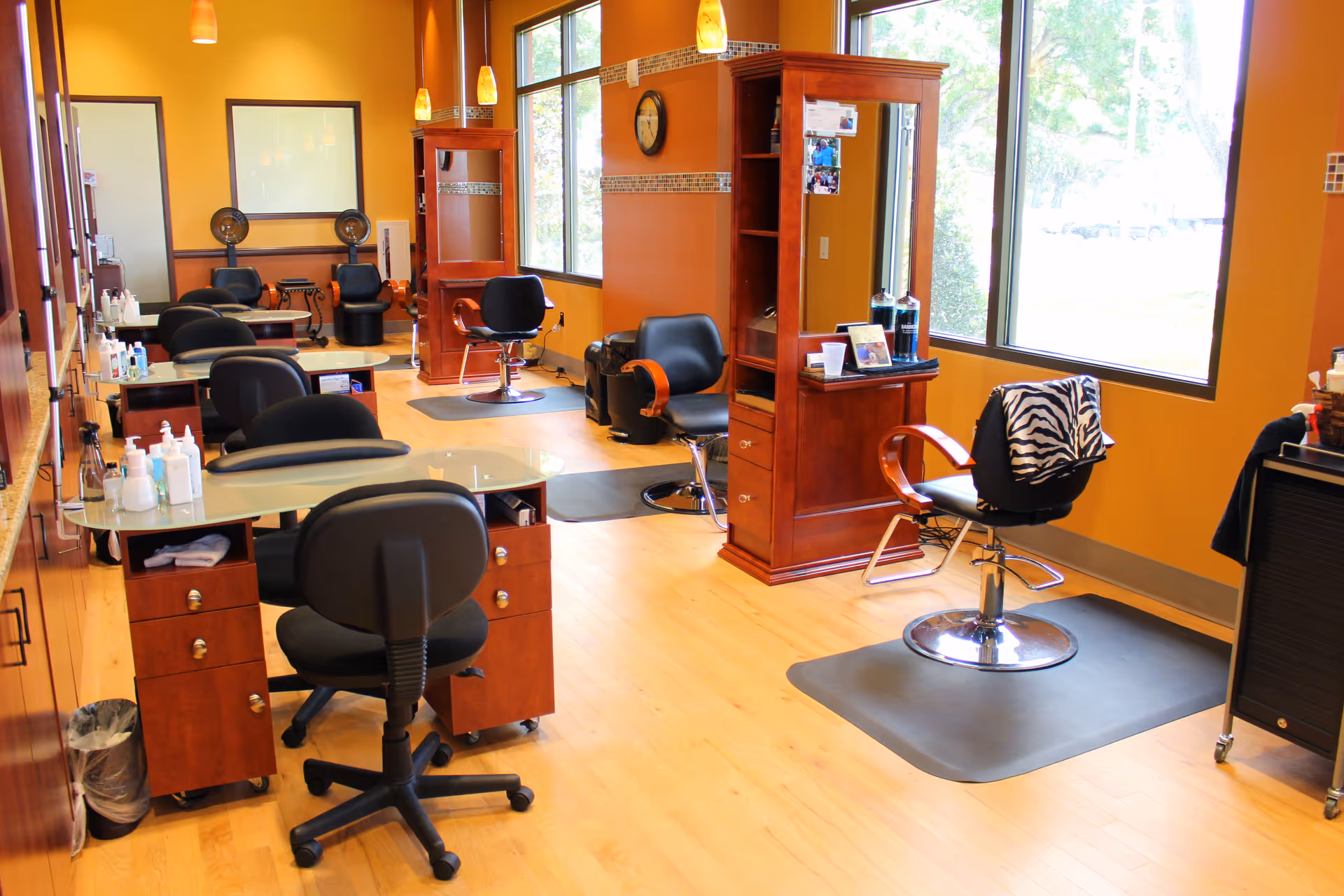 Interior of a salon area with multiple styling stations featuring chairs, mirrors, and wooden cabinetry. The room has large windows letting in natural light, yellow-orange walls, and wooden flooring. Various salon equipment and products are visible on the counters.