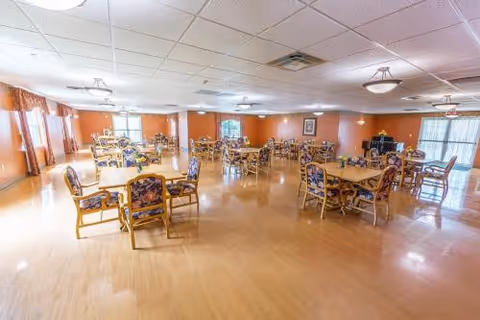 A spacious dining room with multiple wooden tables and chairs arranged throughout the room. The chairs have floral patterned cushions. The room has large windows with curtains, light-colored wooden flooring, and ceiling lights providing illumination. The walls are painted a warm orange color.