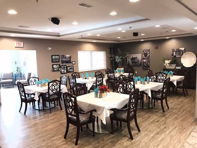 A dining room with multiple square tables covered with white tablecloths and set with teal napkins. Each table is surrounded by dark wooden chairs with intricate back designs. The room has wood flooring, beige walls adorned with framed pictures, and a window letting in natural light. There is a sideboard with decorative items and plants, and an exit door is visible in the background.