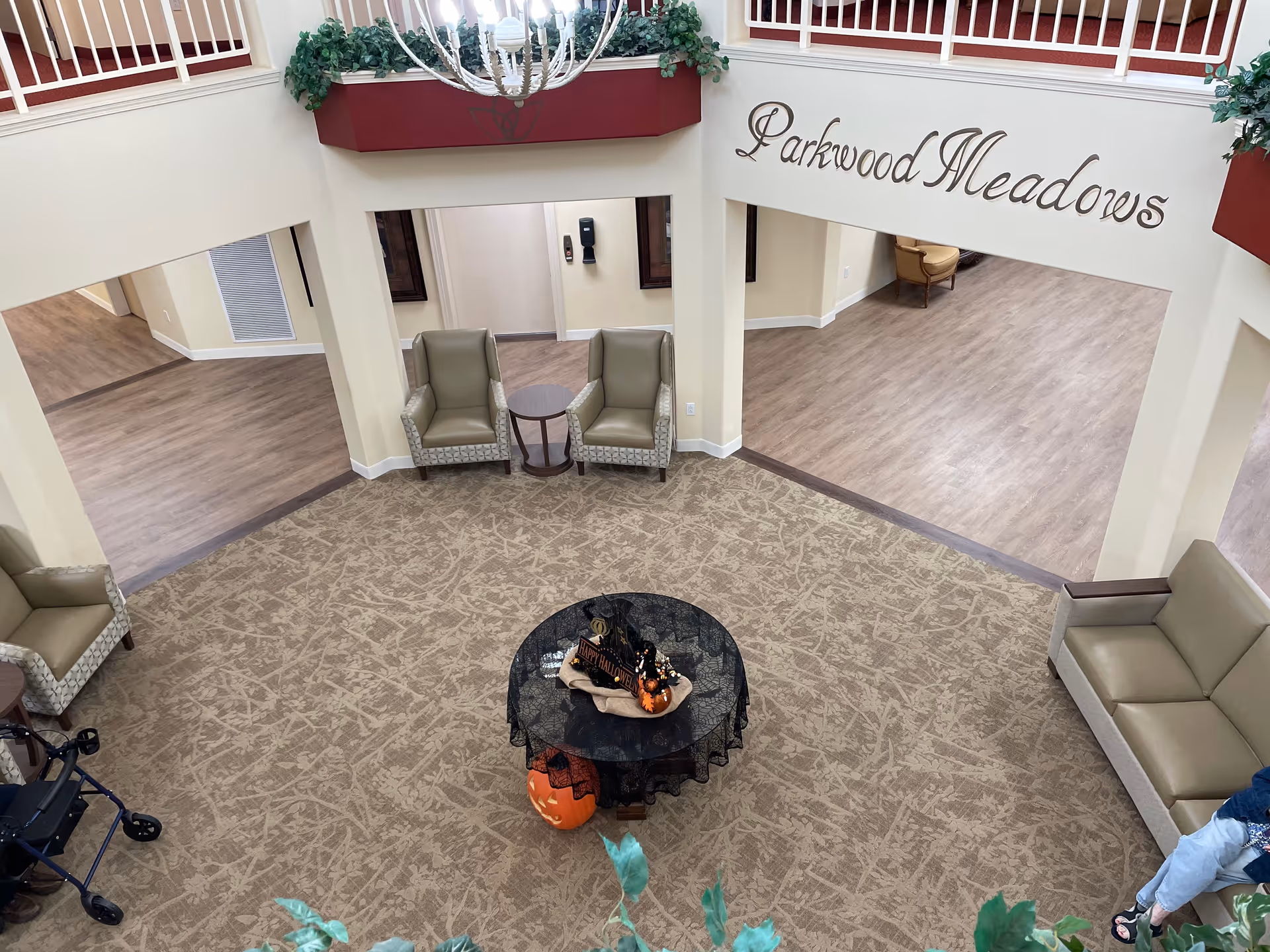 Interior view of a senior living facility common area with beige patterned carpet, several beige armchairs and a sofa arranged around a small round table covered with a black lace cloth and Halloween decorations. The walls are cream-colored with wooden flooring in adjacent rooms. A chandelier hangs from the ceiling, and the words 'Parkwood Meadows' are displayed on the wall above an open hallway.