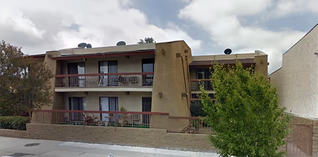 Exterior view of a two-story residential building with balconies on both floors, surrounded by a low wall and some greenery including a tree in the foreground. The sky is partly cloudy.