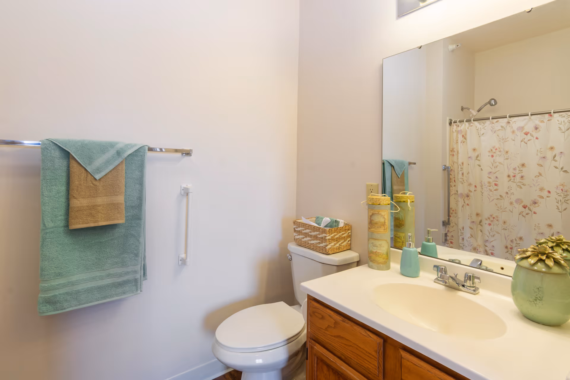 A bathroom with a white toilet, a wooden vanity with a white countertop and sink, a large mirror above the sink, a floral shower curtain, a towel rack with green and beige towels, a basket with towels on the toilet tank, and decorative items on the countertop including a green vase and two cylindrical containers.