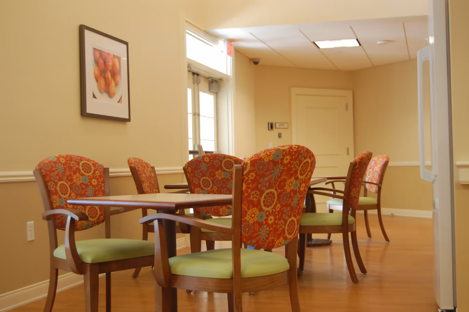 A dining area with wooden tables and chairs featuring green seats and red backs with floral patterns. The room has beige walls, a framed picture of apples on the wall, and a door with windows letting in natural light.