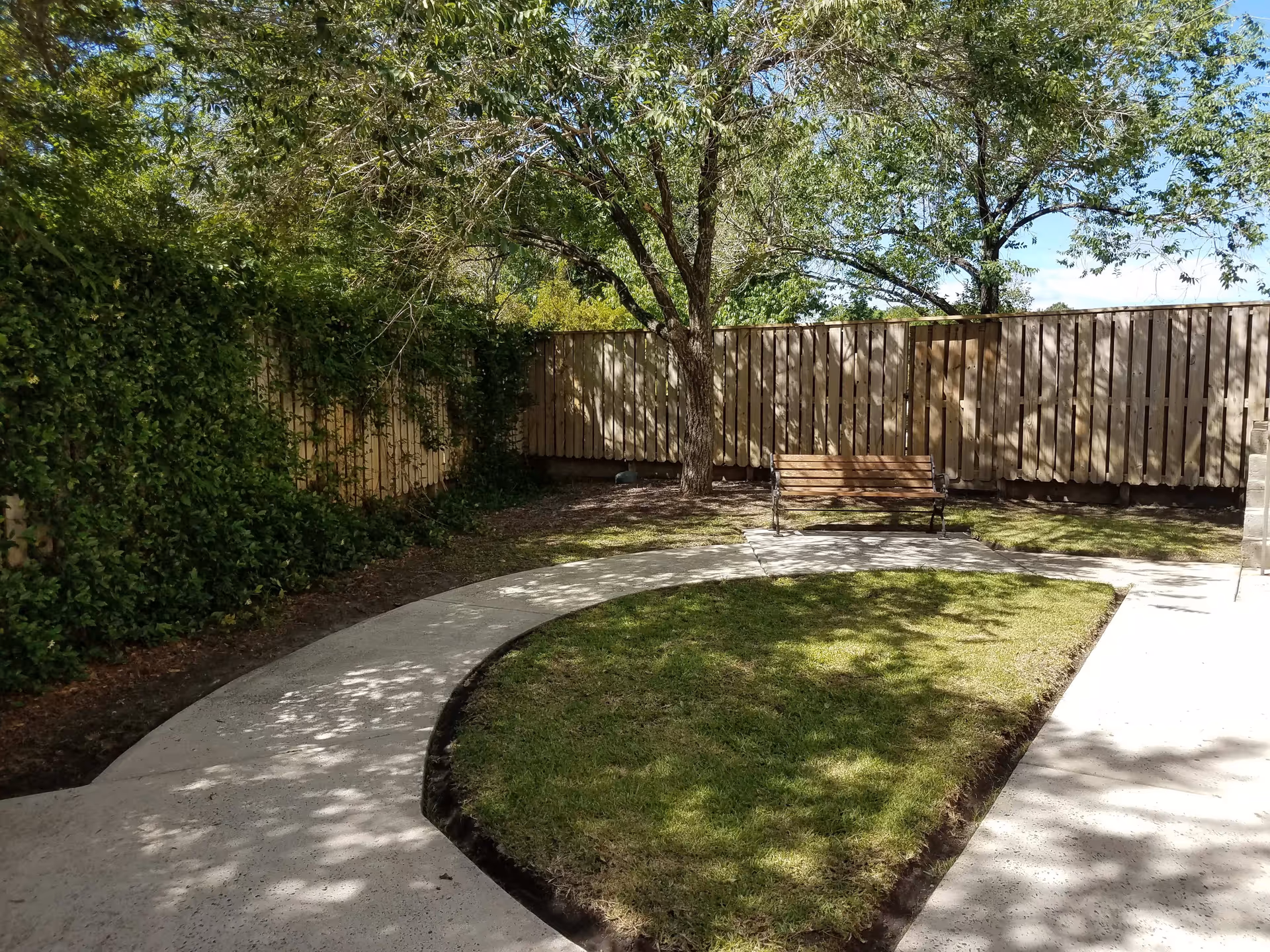 A peaceful outdoor garden area with a curved concrete pathway, a wooden bench under a tree, green grass, and a wooden fence surrounding the space.