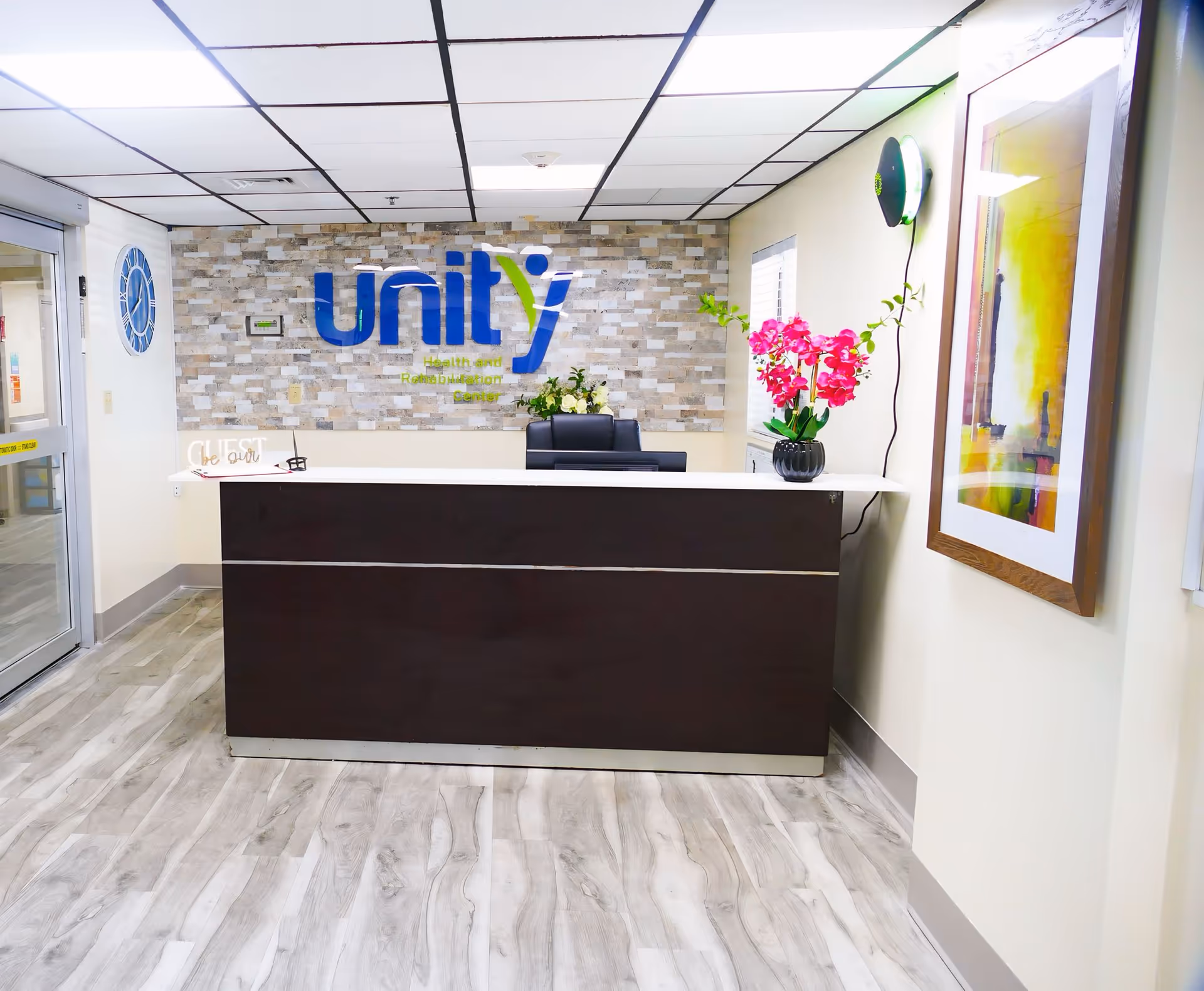 Reception area of Unity Health and Rehabilitation Center with a modern dark wood front desk, a decorative stone wall behind with the center's logo, a vase of pink flowers on the desk, a framed colorful abstract painting on the right wall, and a clock on the left wall near the entrance door.