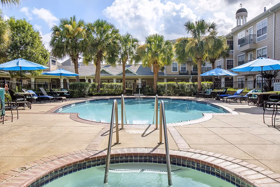 Outdoor swimming pool area at Brookdale First Colony with palm trees, blue umbrellas, lounge chairs, and a hot tub in the foreground. Residential buildings surround the pool under a partly cloudy sky.