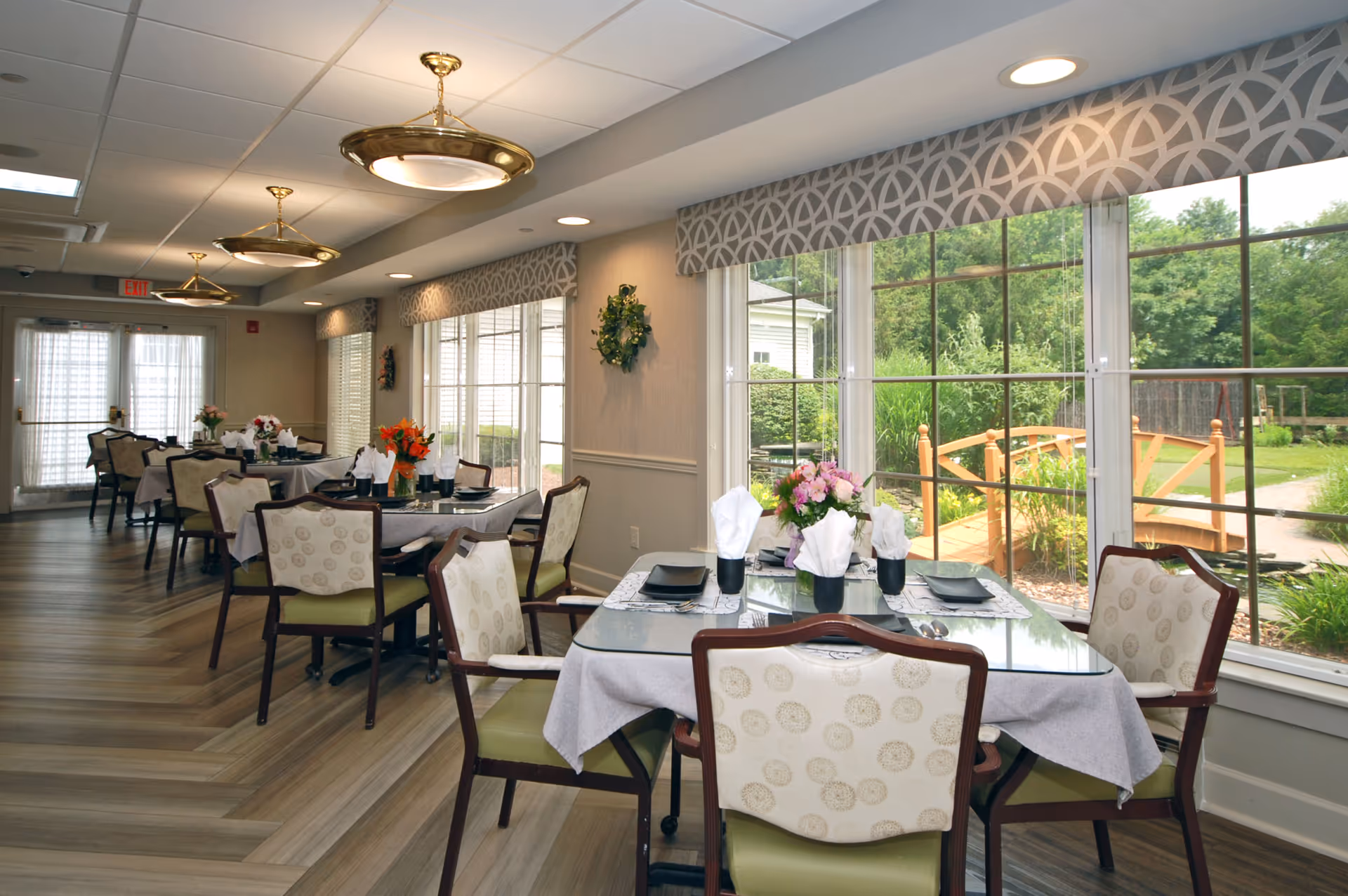 A bright dining room with several tables set for meals, each with white napkins, black plates, and floral centerpieces. Large windows on one side provide a view of a garden with a wooden bridge and greenery outside. The room features patterned valances, wooden chairs with cushioned seats, and modern ceiling lights.