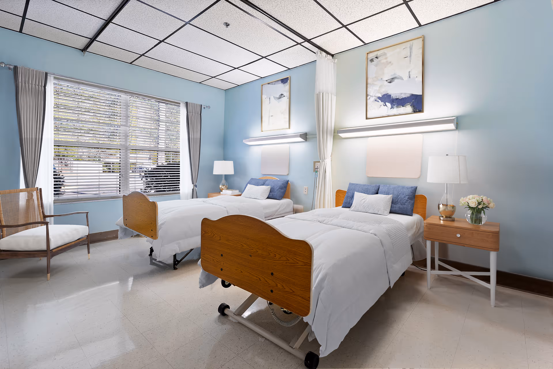 A bright and clean bedroom in a senior living facility with two single beds made with white linens and blue pillows. Each bed has a wooden headboard and footboard, and there is a nightstand with a lamp and a vase of flowers between the beds. The walls are painted light blue and decorated with abstract artwork. A large window with blinds and curtains lets in natural light, and a wooden chair with a white cushion is placed near the window.