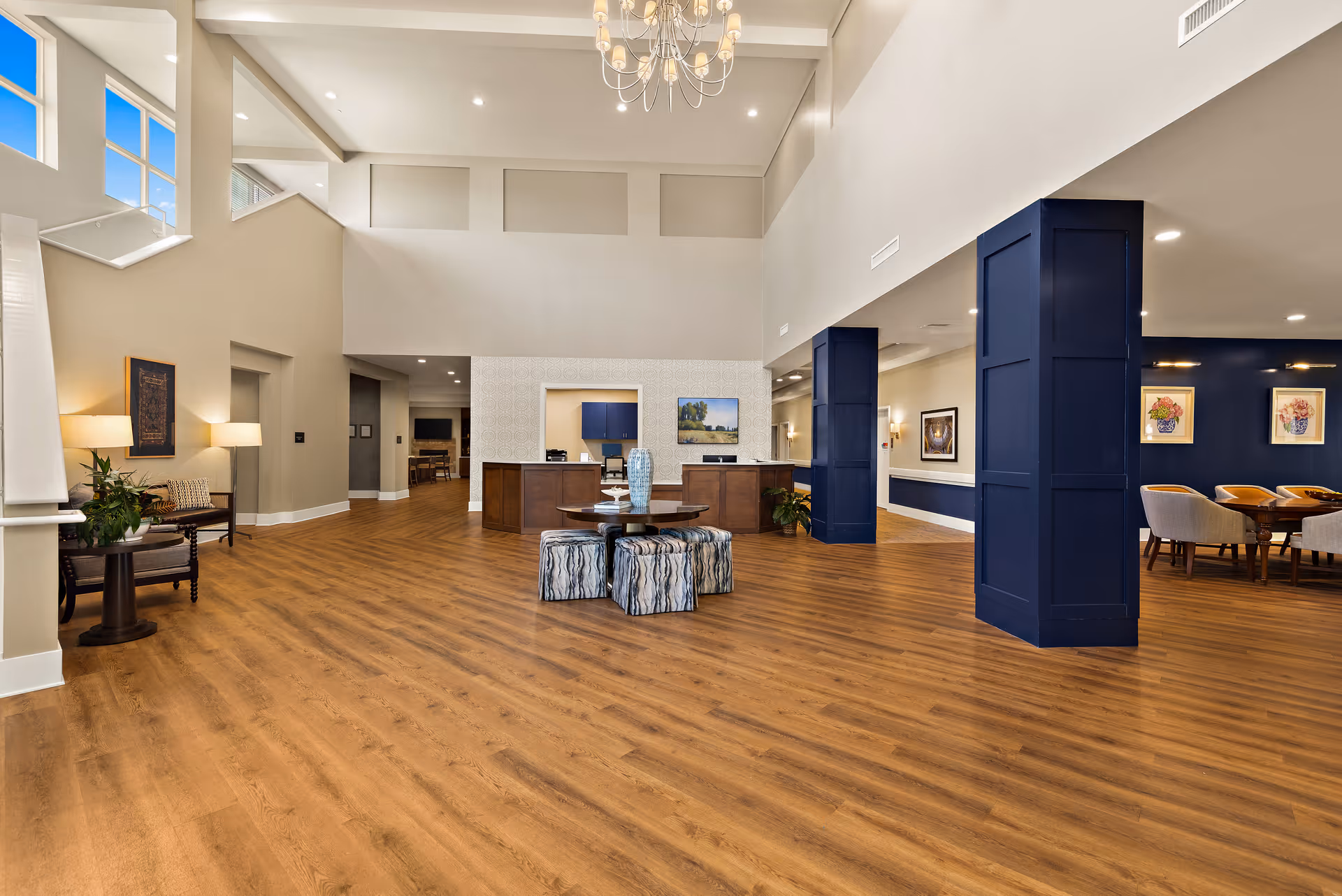 Spacious and well-lit interior common area with wooden flooring, a round table with decorative stools in the center, blue accent columns, and a reception desk in the background. There are framed artworks on the walls and a chandelier hanging from the ceiling.