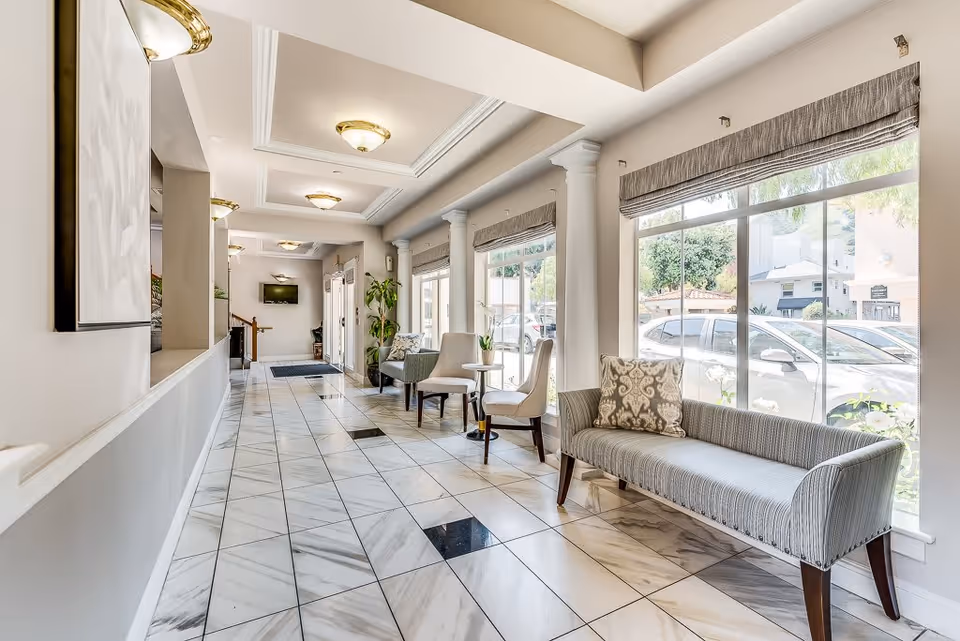 Bright and spacious hallway in a senior living facility with large windows on the right side letting in natural light. The floor is tiled with a white and gray marble pattern with some black tiles. Along the windows are several seating options including a striped bench with a decorative pillow and two white chairs with a small round table between them. The ceiling has recessed lighting fixtures and decorative molding. There are columns along the window side and a flat-screen TV mounted on the far wall near the entrance door.